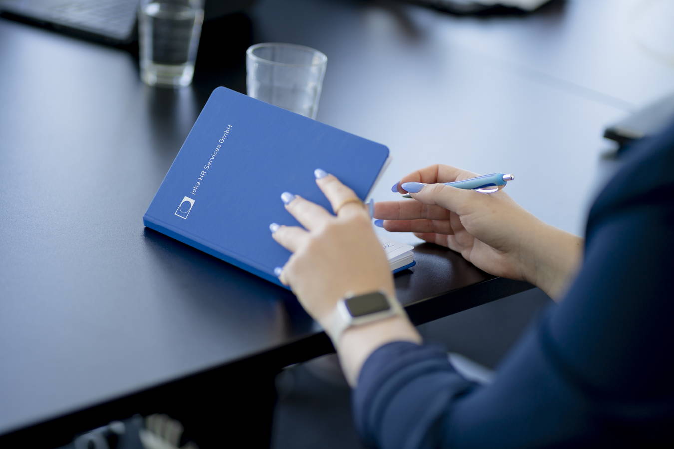 Person hält einen blauen Notizblock mit der Aufschrift „Joka HR Services GmbH“ und einen Stift am Tisch