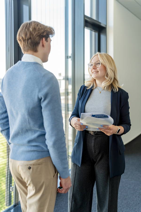 Zwei Personen unterhalten sich in einem hellen Büroflur; die Frau hält Unterlagen in der Hand