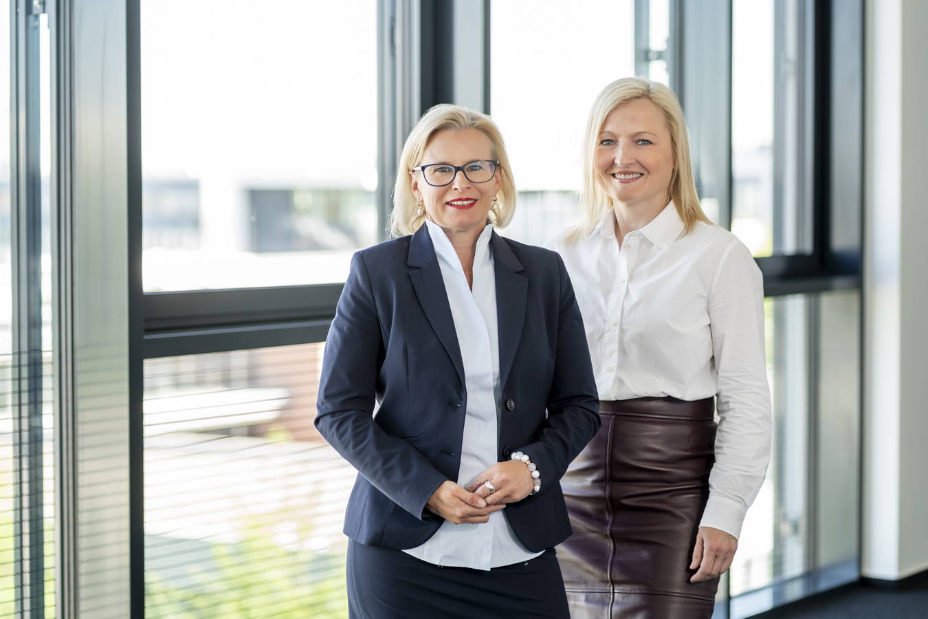Zwei lächelnde Geschäftsfrauen stehen vor großen Fenstern in einem Büro