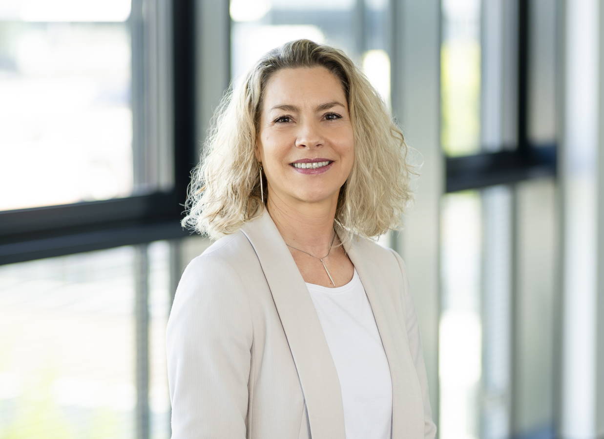 Porträt einer lächelnden Frau in heller Businesskleidung vor großen Fenstern