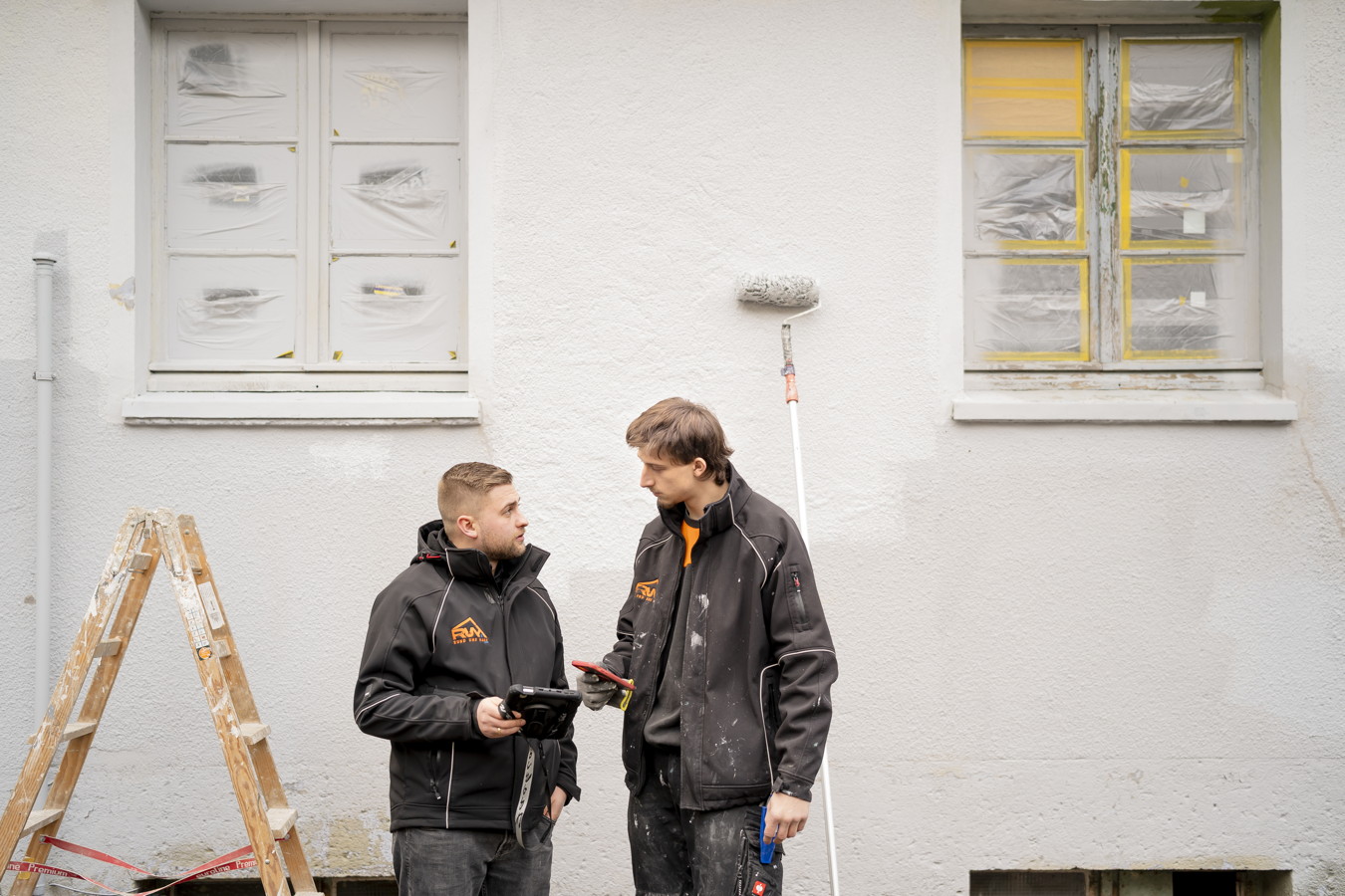 Zwei Handwerker besprechen die Fassadenrenovierung vor einem Haus mit abgeklebtfenstern