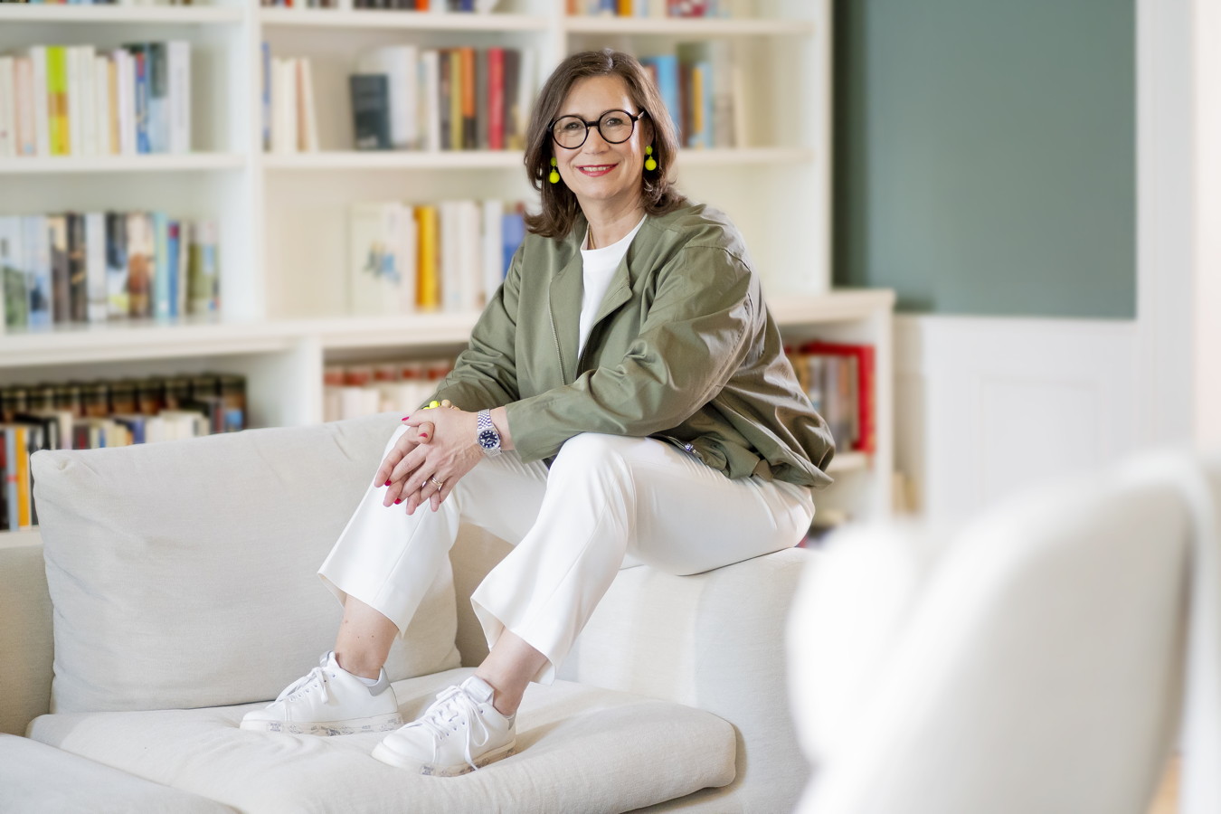 Businessfotos Frau sitzt lächelnd auf einem hellen Sofa vor einem Bücherregal