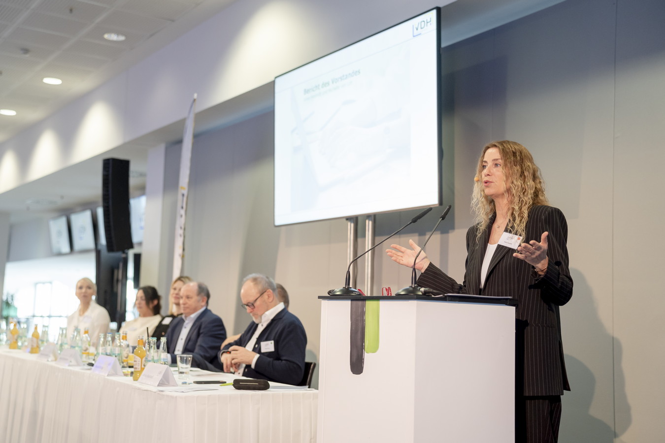 Rednerin spricht an einem Podium bei einer Konferenz, daneben sitzen mehrere Teilnehmende am Tisch