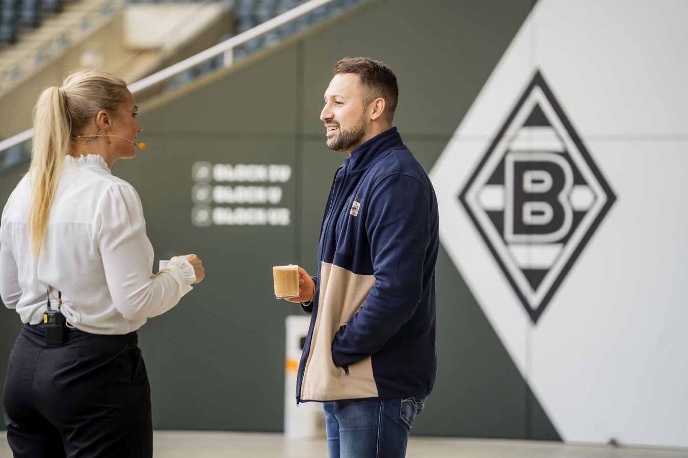 Zwei Personen unterhalten sich und halten Kaffeetassen vor einer Borussia-Mönchengladbach-Wand