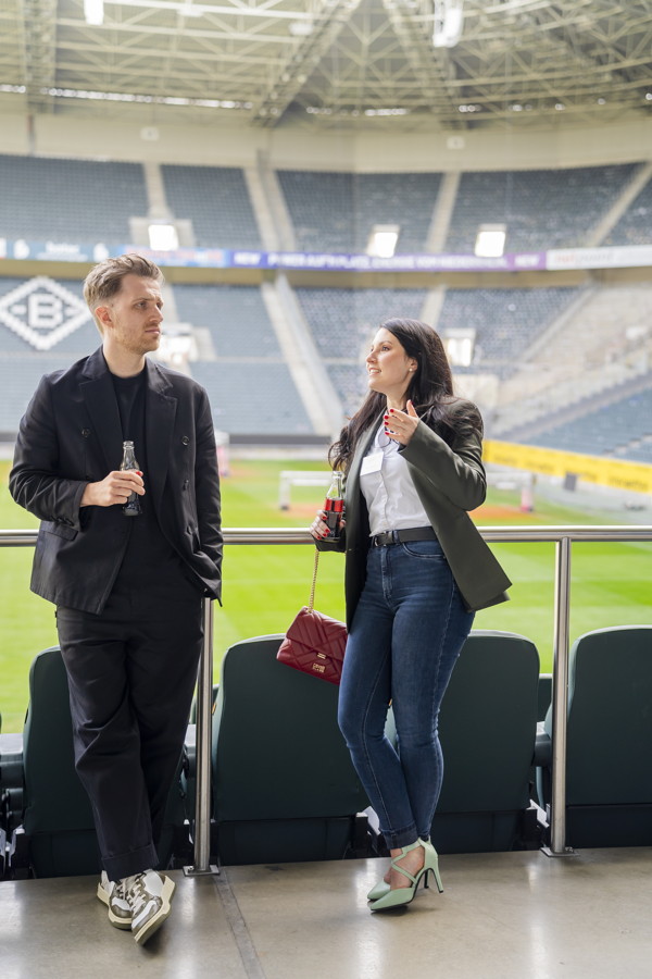 Zwei Personen unterhalten sich auf einer Stadiontribüne und halten Getränke in der Hand