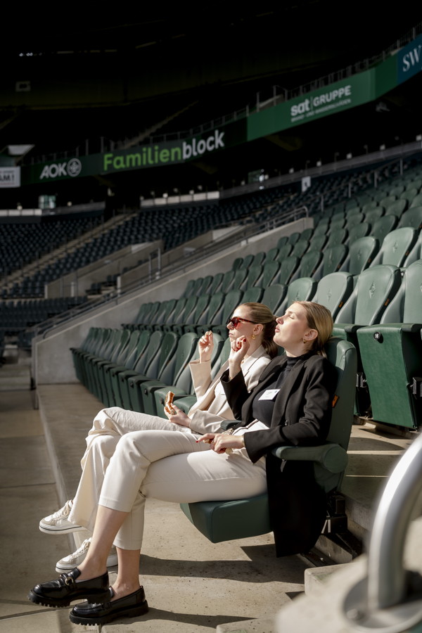 Zwei Frauen sitzen in einem leeren Stadion auf grünen Sitzen und essen Snacks im Familienblock