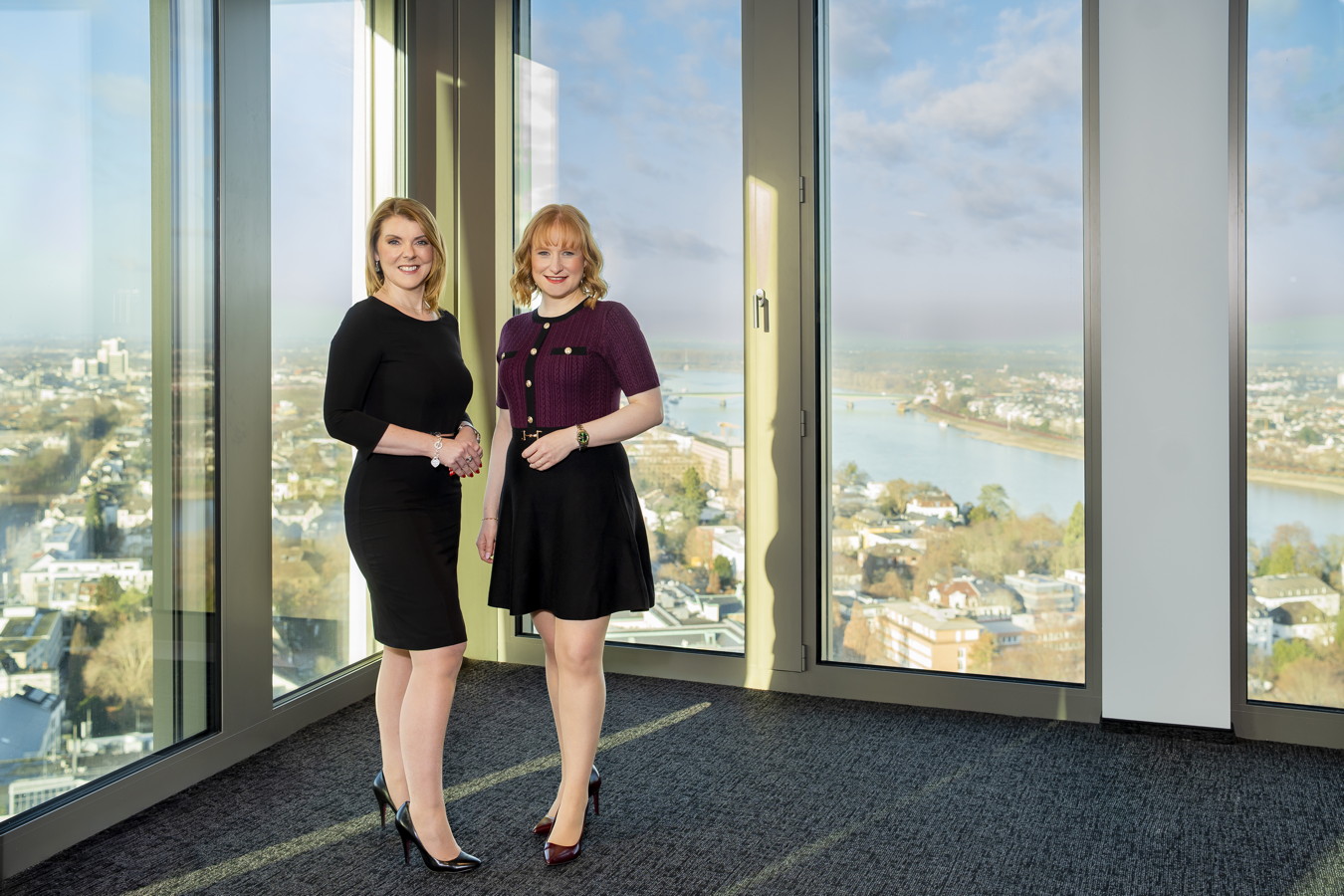 Zwei Frauen stehen in einem Büro vor großen Fenstern mit Blick auf eine Stadt und einen Fluss