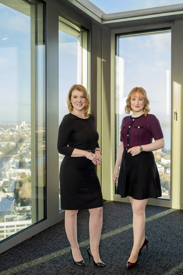 Zwei Frauen stehen in einem hellen Raum vor großen Fenstern mit Stadtblick