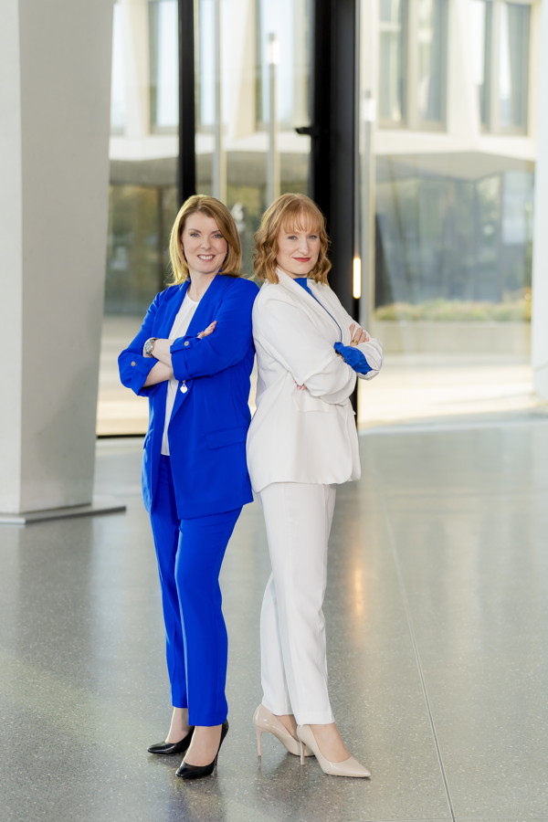 Zwei Frauen in blauen und weißen Anzügen stehen Rücken an Rücken mit verschränkten Armen in einem hellen Atrium