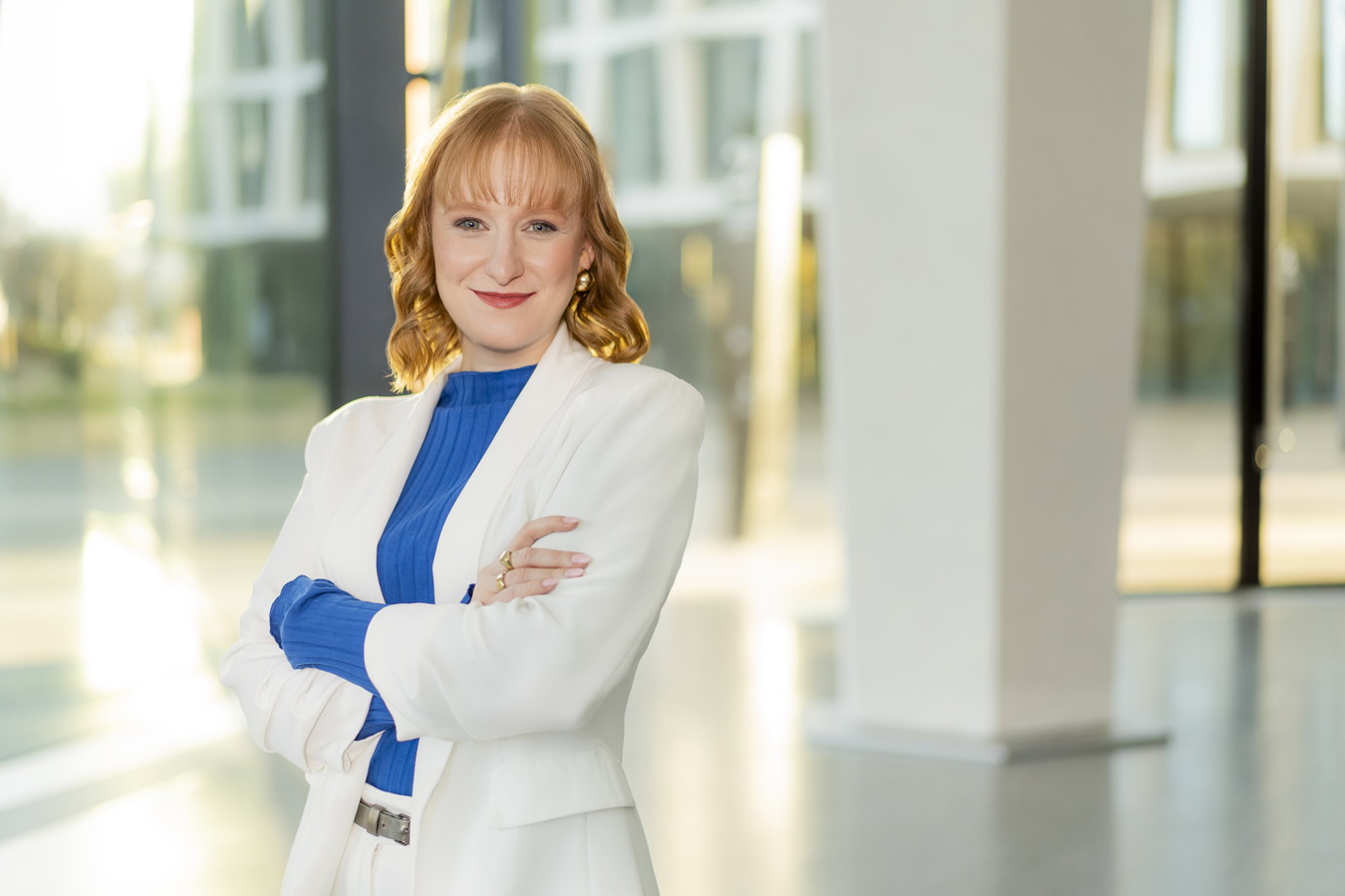 Porträt einer Frau in weißem Blazer und blauem Oberteil mit verschränkten Armen in einem hellen Gebäude