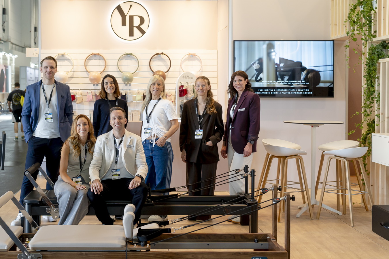 Sieben Personen posieren an einem Messestand mit Pilates-Reformer und YR-Logo